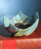 A handmade ceramic lotus-shaped bowl with a glossy green and blue glaze, resting on a red vintage book against a blue background.