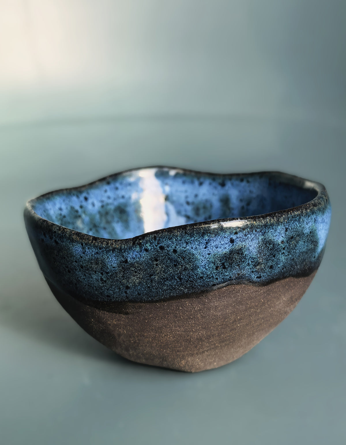 A handmade ceramic bowl with a wavy, organic rim, featuring a glossy textured blue glaze on the top half and a matte, earthy brown clay finish on the bottom half, set against a soft blue-gray background.
