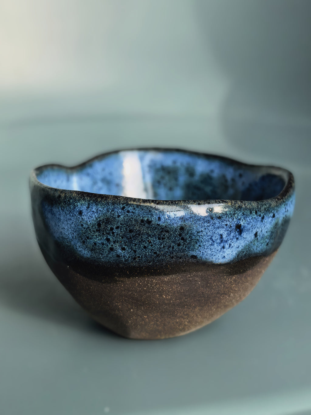 A handmade ceramic bowl with a wavy rim, featuring a glossy blue and black speckled glaze on the upper half and a matte, textured brown base on the lower half, set against a soft, muted blue-gray background.