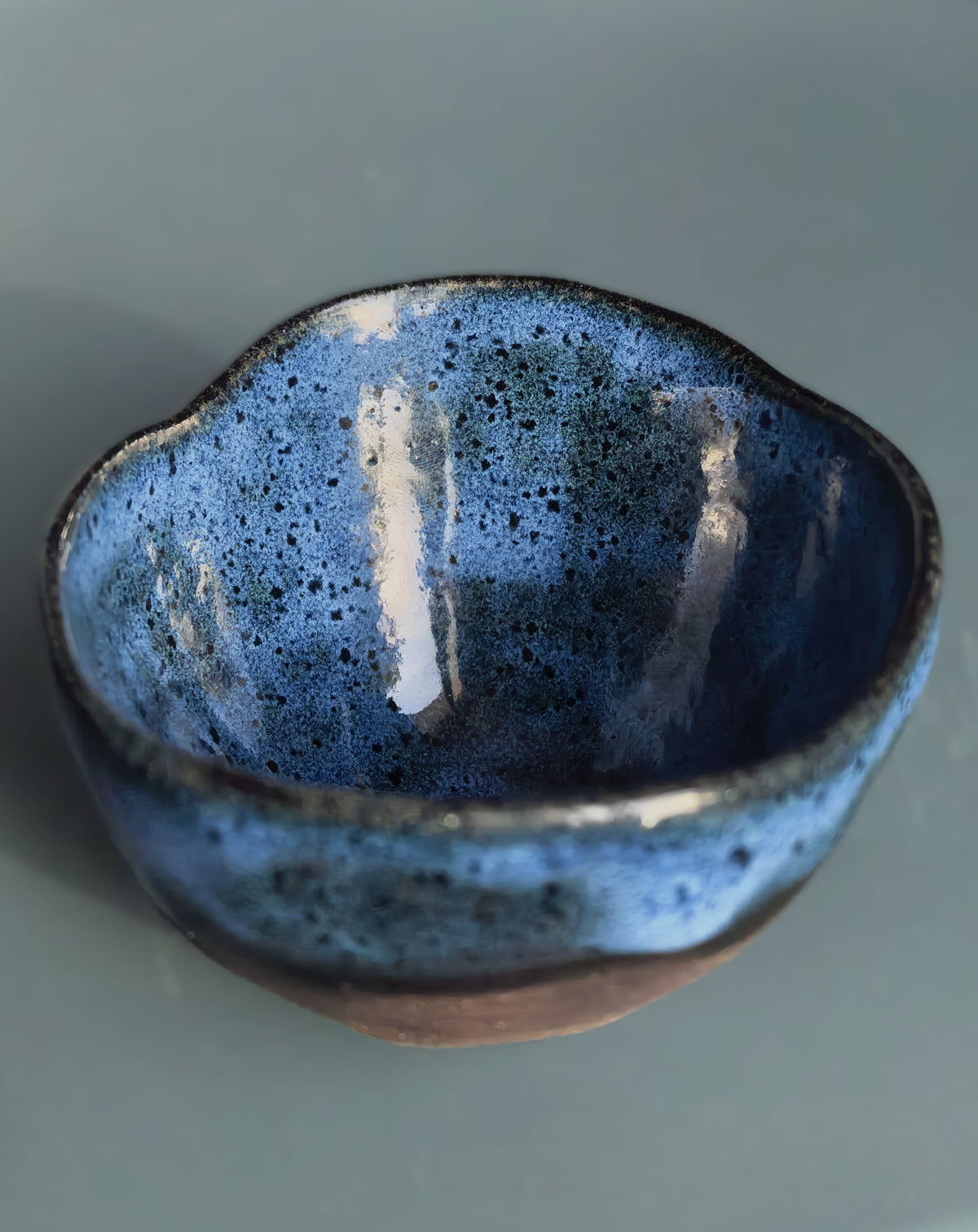 A handmade ceramic tea bowl with a textured, speckled blue and black glaze and a rustic brown base, shown on a smooth gray surface.