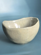 A handmade ceramic bowl with a textured, crackled glaze in a light beige or off-white color, set against a soft blue background.