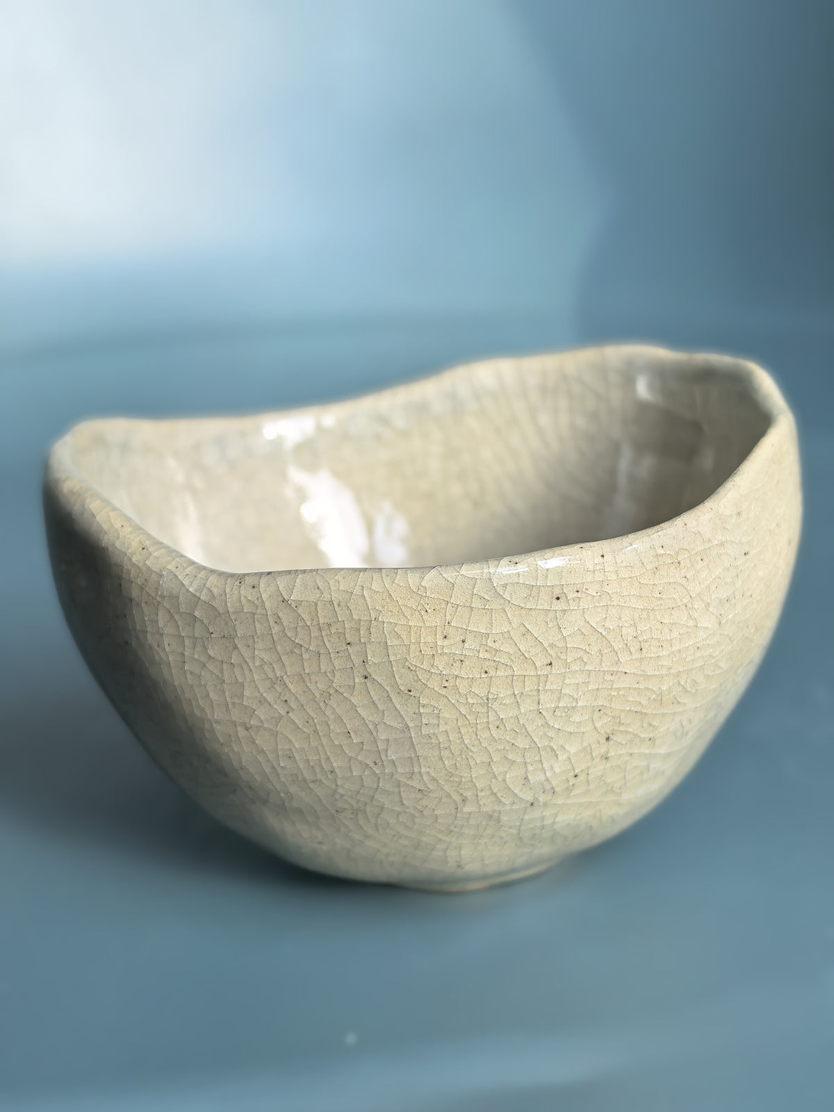 A handmade ceramic bowl with a textured, crackled glaze in a light beige or off-white color, set against a soft blue background.
