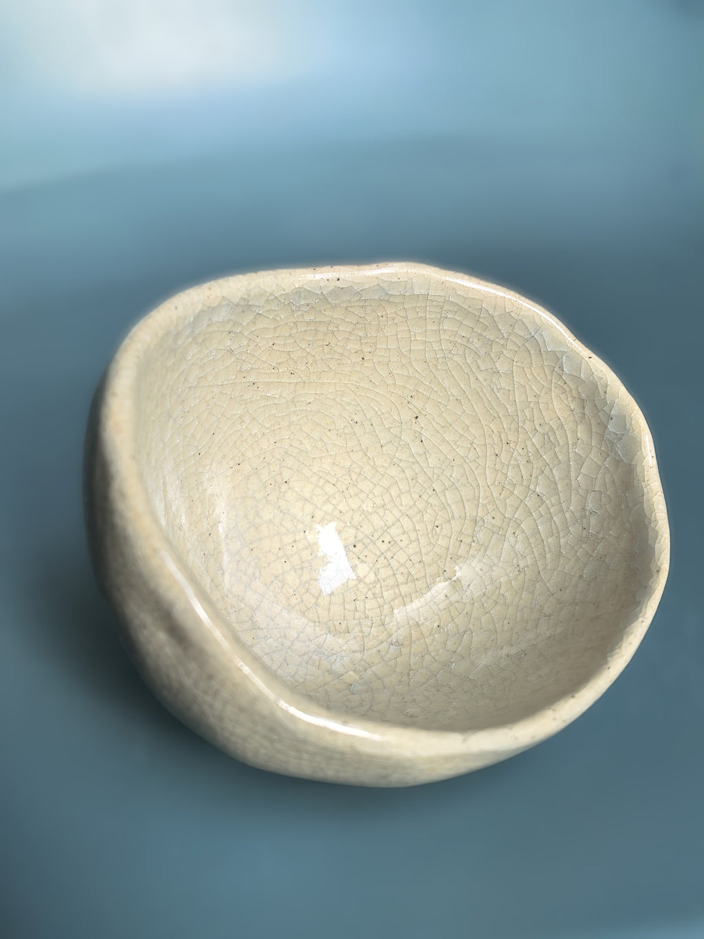 An organic-shaped ceramic bowl with a glossy, crackled glaze in a light beige or off-white color, set against a smooth, solid blue background.