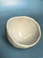 An organic-shaped ceramic bowl with a glossy, crackled glaze in a light beige or off-white color, set against a smooth, solid blue background.