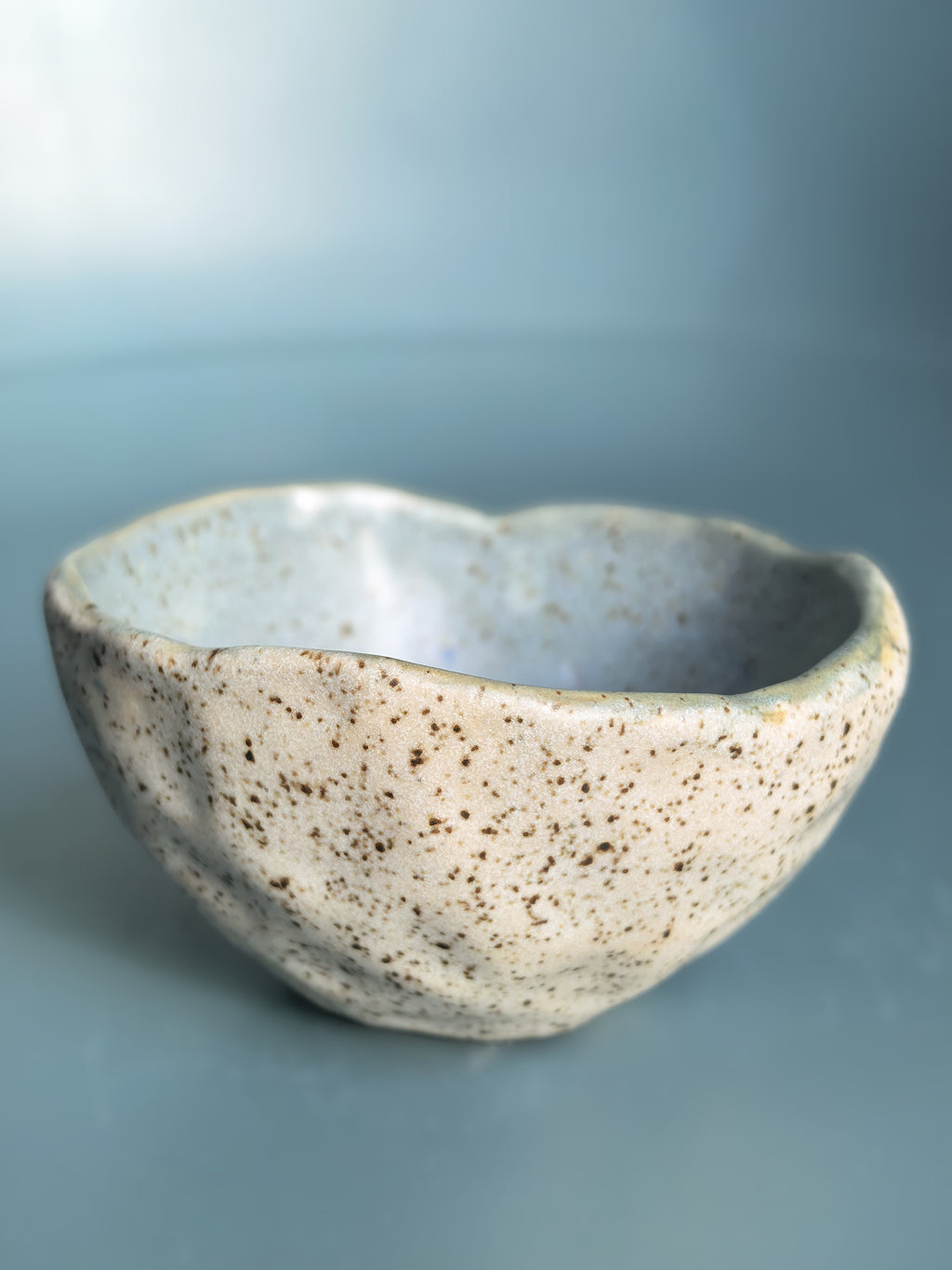 A handmade ceramic bowl with a wavy, organic rim and a speckled glaze, shown on a smooth blue-gray background.
