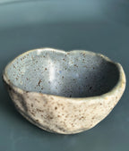 A handmade ceramic bowl with a textured, organic shape, featuring a speckled beige exterior and a smooth, slate-blue interior, set against a solid dark teal background.
