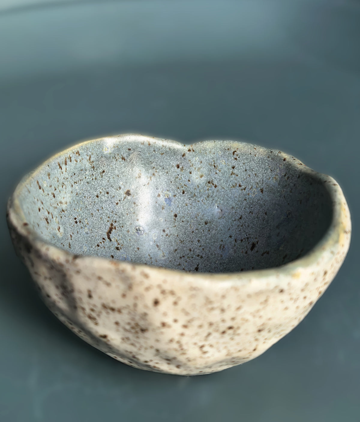 A handmade ceramic bowl with a textured, organic shape, featuring a speckled beige exterior and a smooth, slate-blue interior, set against a solid dark teal background.