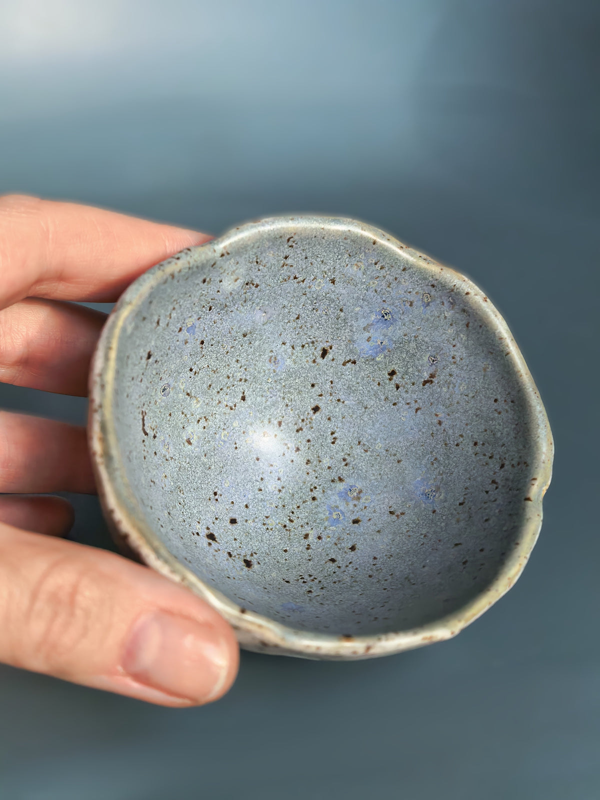 A handmade ceramic bowl with a speckled slate-blue interior and a rough, natural beige rim, held by a hand against a soft blue background.