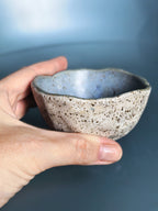 A handmade ceramic bowl with a speckled beige exterior and a light blue glazed interior, held in a person's hand against a soft blue background.