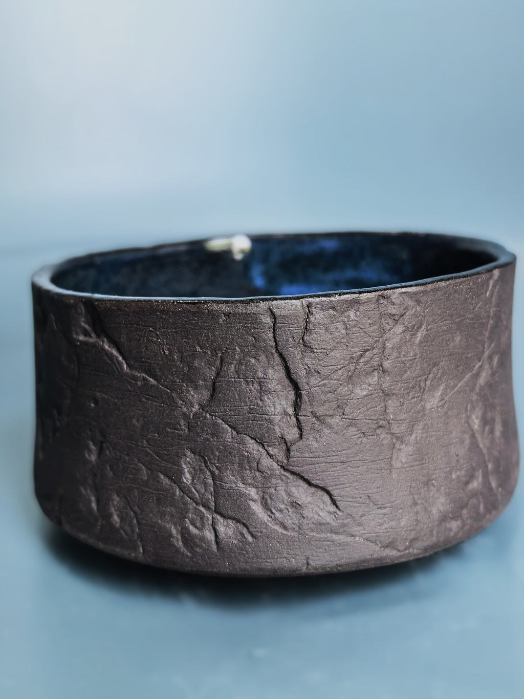 A wide, cylindrical dark brown textured leather or faux-leather ring holder with a dark blue velvet interior, shown on a light blue background.