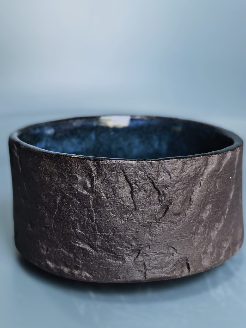 A wide, textured ceramic bowl with a dark brown, earthy exterior and a glossy dark blue interior, set against a soft light blue background.