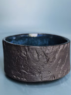 A wide, textured ceramic bowl with a dark brown, earthy exterior and a glossy dark blue interior, set against a soft light blue background.