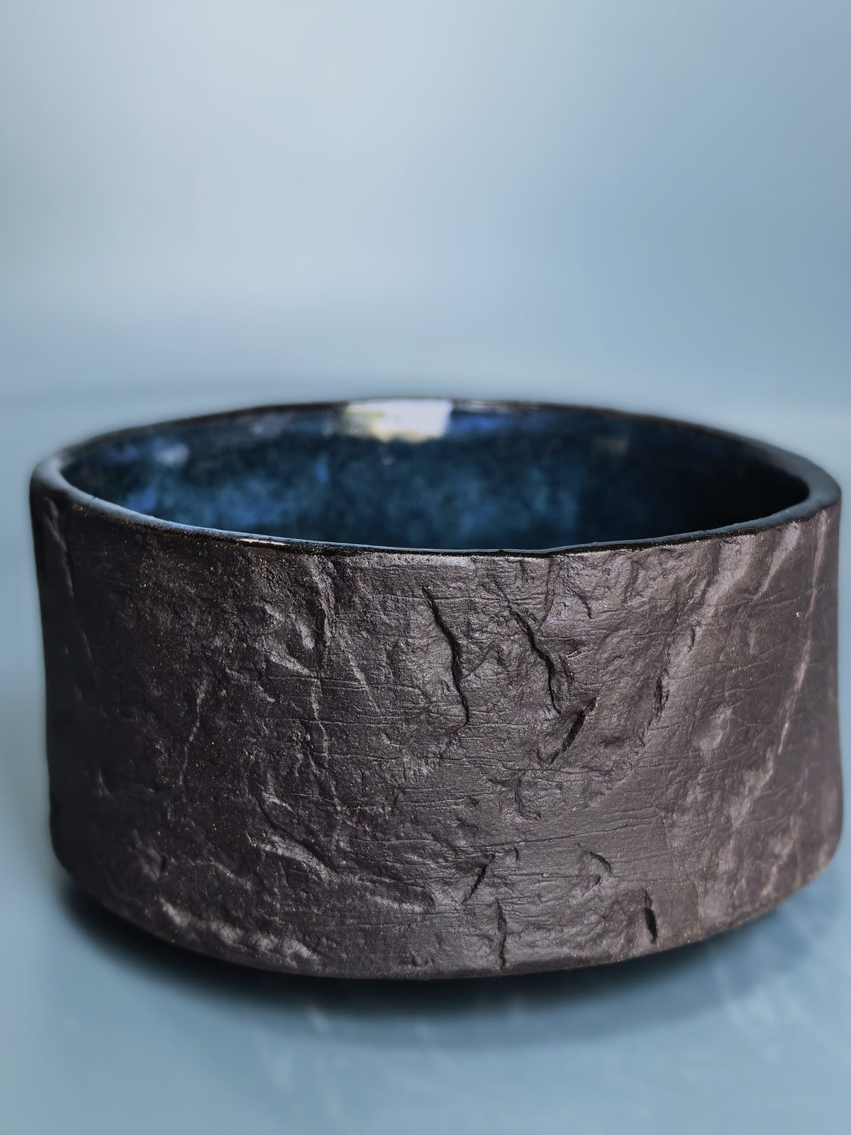 A wide, textured ceramic bowl with a dark brown, earthy exterior and a glossy dark blue interior, set against a soft light blue background.