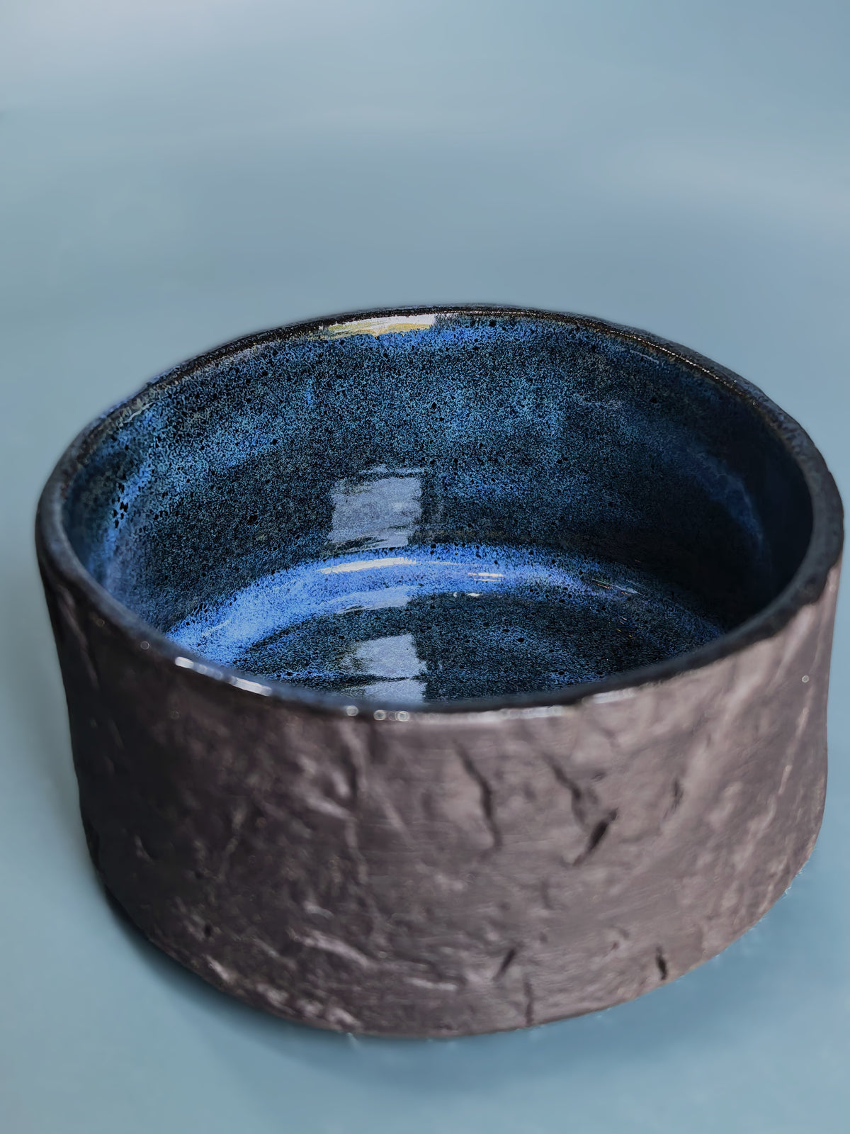 A handmade ceramic bowl with a textured, dark brown exterior and a glossy, speckled blue interior, set against a solid light blue background.
