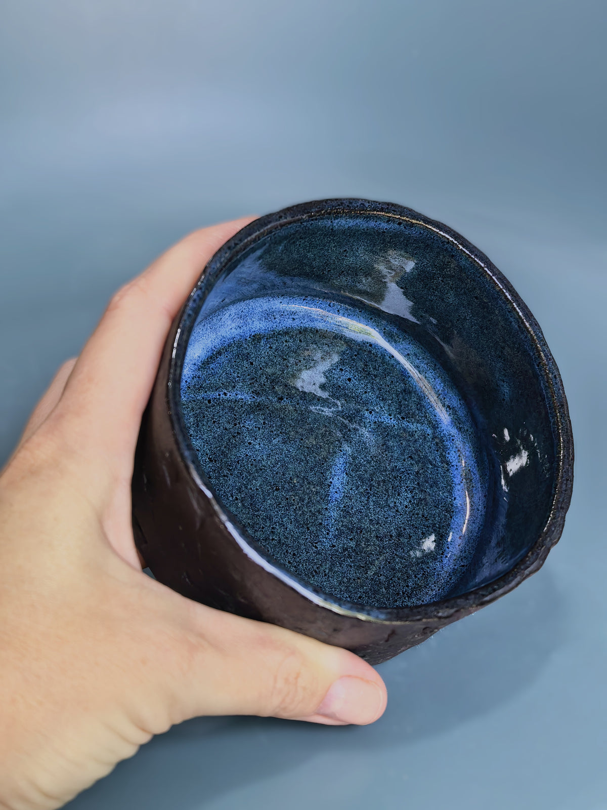 A hand holding a handmade ceramic bowl with a textured dark brown exterior and a glossy, speckled deep blue interior, set against a solid blue-gray background.