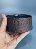 A hand holding a wide, textured dark brown ceramic bowl with a glossy dark blue interior, set against a soft blue-gray background.