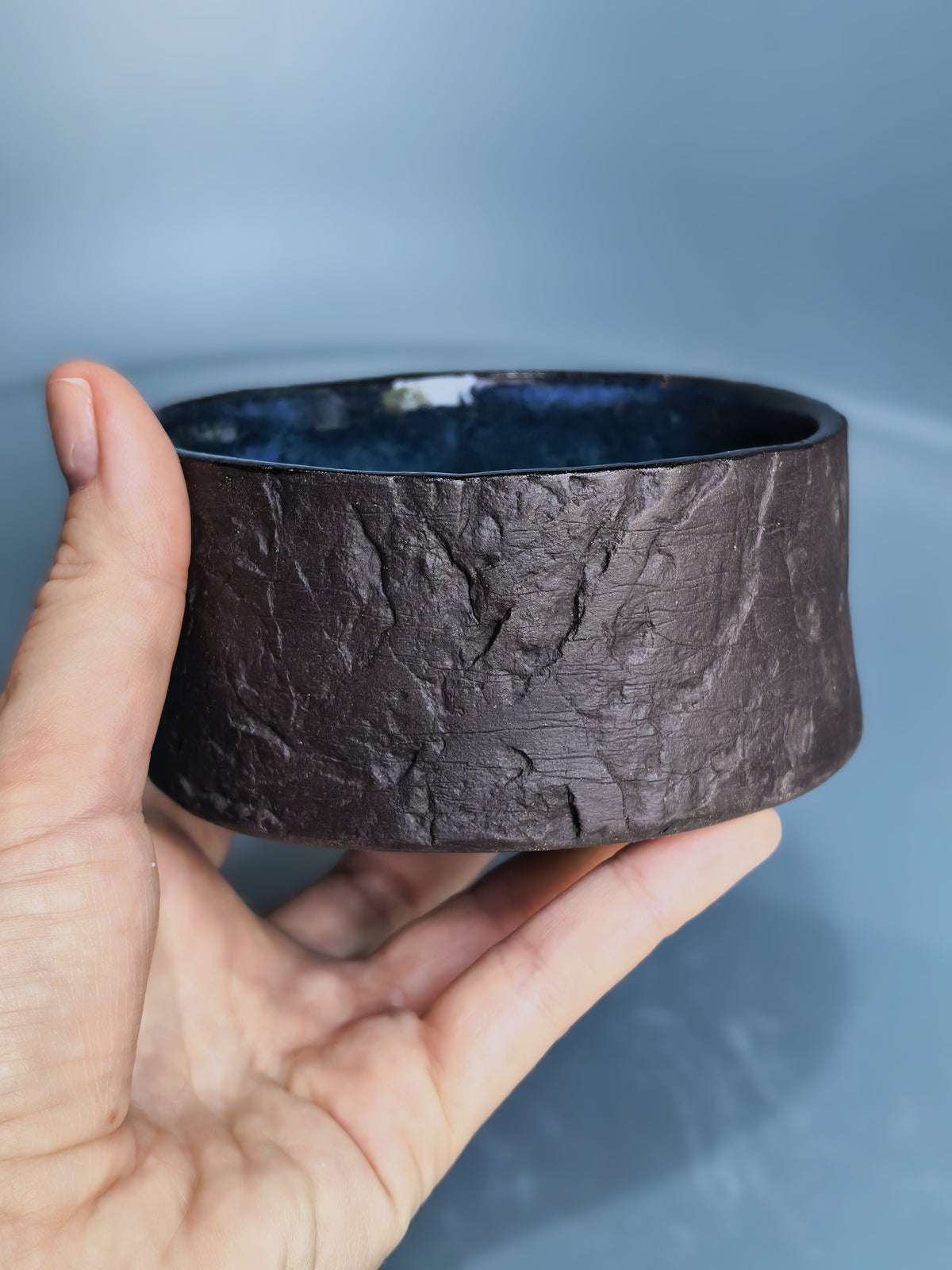 A hand holding a wide, textured dark brown ceramic bowl with a glossy dark blue interior, set against a soft blue-gray background.