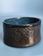 A handmade ceramic bowl with a textured, metallic bronze exterior featuring embossed floral patterns and a speckled dark blue-gray glazed interior, set against a soft light blue background.