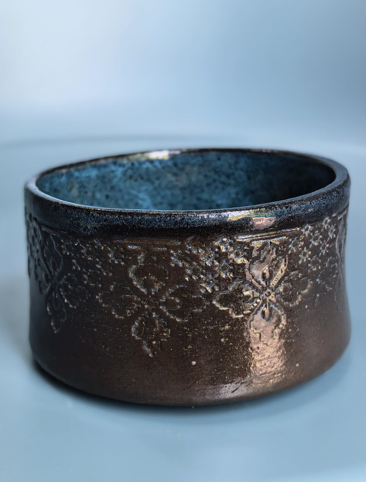 A handmade ceramic bowl with a textured, metallic bronze exterior featuring embossed floral patterns and a speckled dark blue-gray glazed interior, set against a soft light blue background.