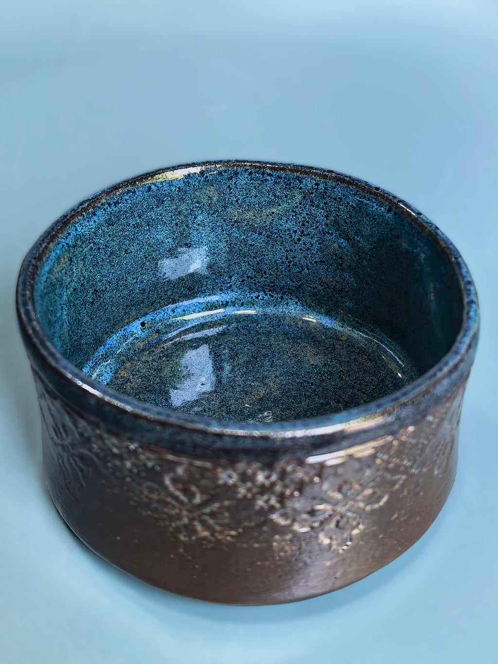 A handmade ceramic bowl with a textured, speckled glaze transitioning from deep blue on the inside to a dark brown with metallic gold flecks on the outside, set against a solid light blue background.