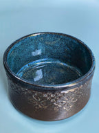 A handmade ceramic bowl with a textured, speckled glaze transitioning from deep blue on the inside to a dark brown with metallic gold flecks on the outside, set against a solid light blue background.