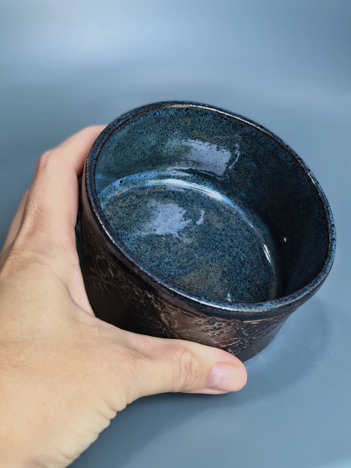 A hand holding a handmade ceramic bowl with a textured exterior and a glossy, speckled dark blue glaze interior, set against a solid blue-gray background.