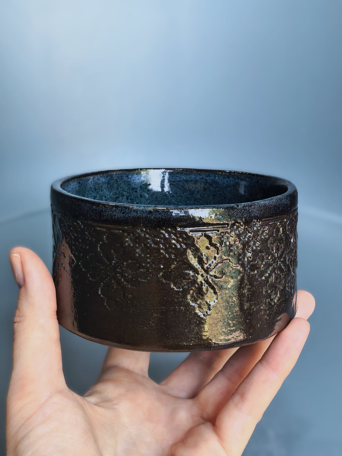 A handmade dark brown ceramic bowl with a textured, metallic gold leaf pattern on the exterior, held in a person's hand against a soft blue-gray background.