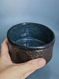 A hand holding a small handmade ceramic bowl with a dark blue speckled interior and a textured dark brown exterior, set against a soft blue-gray background.