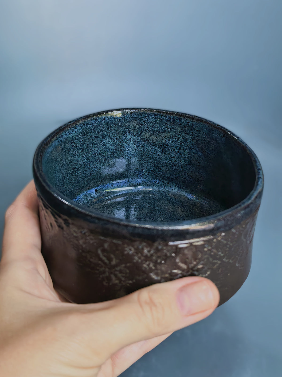 A hand holding a small handmade ceramic bowl with a dark blue speckled interior and a textured dark brown exterior, set against a soft blue-gray background.