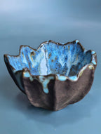 A handmade ceramic bowl with a wavy, scalloped rim, featuring a glossy blue glazed interior and a dark brown textured exterior with light blue accents, set against a soft blue background.
