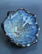 A handmade ceramic bowl with a textured, organic shape and a blue, speckled glaze, photographed on a smooth dark gray surface.