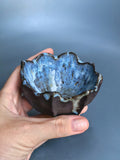 A hand holding a small handmade ceramic bowl with a dark brown exterior and a blue speckled glazed interior, set against a soft blue-gray background.