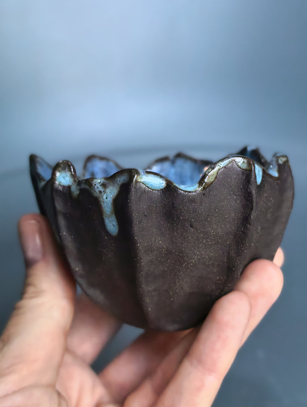 A handmade ceramic bowl with a dark brown exterior and a blue glazed interior, held in a person's hand against a soft blue-gray background.