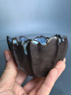A handmade ceramic bowl with a dark brown exterior and a blue glazed interior, held in a person's hand against a soft blue-gray background.
