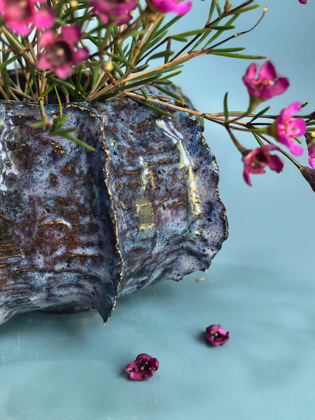 A textured metallic planter with a hammered, oxidized finish in silver and dark gray tones, accented with gold leaf details along the edge. It is filled with green foliage and small pink flowers, set against a smooth light blue background.