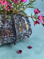 A textured metallic planter with a hammered, oxidized finish in silver and dark gray tones, accented with gold leaf details along the edge. It is filled with green foliage and small pink flowers, set against a smooth light blue background.