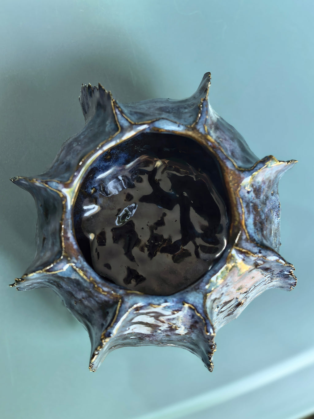 A handmade ceramic bowl with a starburst or sunflower-like rim, featuring a glossy black interior and textured, speckled gray and gold-toned exterior glaze, shown from a top-down perspective on a light blue-green background.