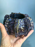 A handmade ceramic planter with a highly textured, organic shape, held in a person's hand against a soft blue-gray gradient background.