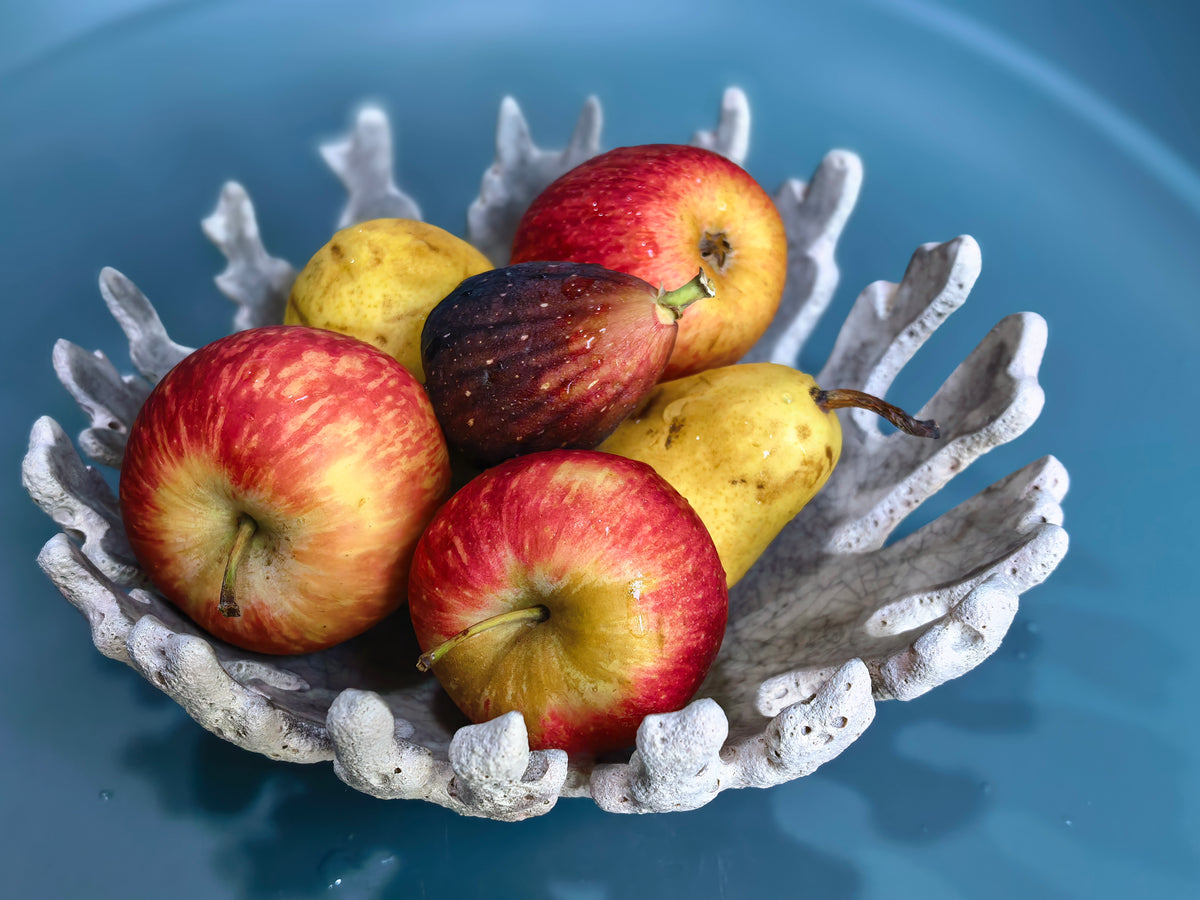 A decorative white coral-shaped bowl filled with red apples, yellow pears, and a dark fig, set against a smooth blue background.