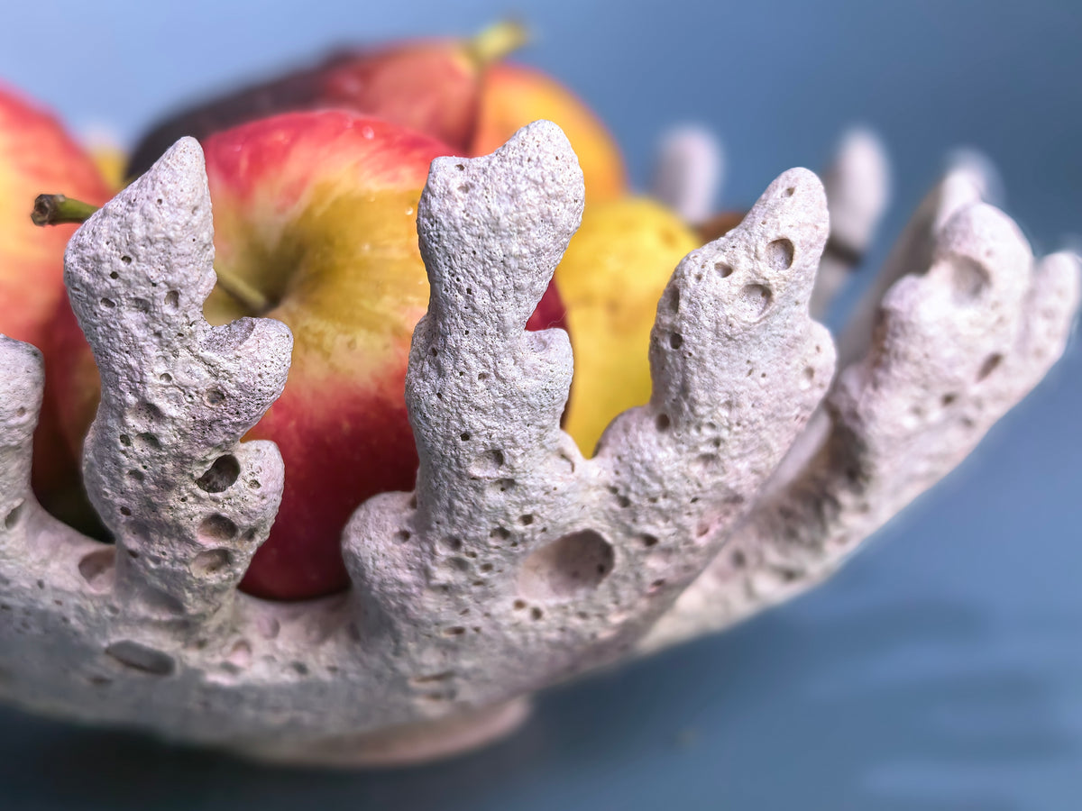 A close-up of a textured, coral-like white bowl filled with red and yellow apples, set against a soft blue background.