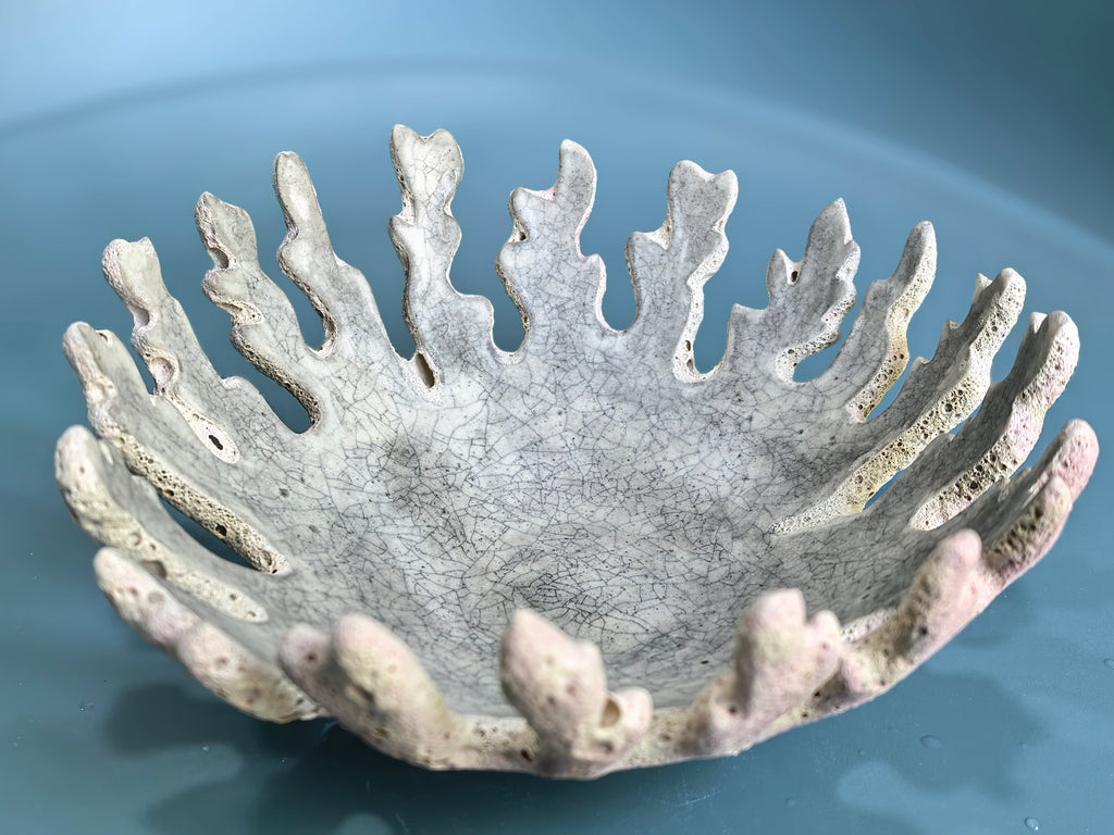 A decorative ceramic bowl with a coral or urchin-like fluted rim, shown on a smooth blue surface. The bowl has a textured, crackled glaze in light gray and off-white tones with subtle pinkish-beige shading along the outer edges.
