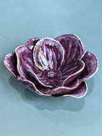 A sculptural ceramic flower bowl in deep purple with glossy highlights and a white glazed edge, set against a solid teal-blue background.