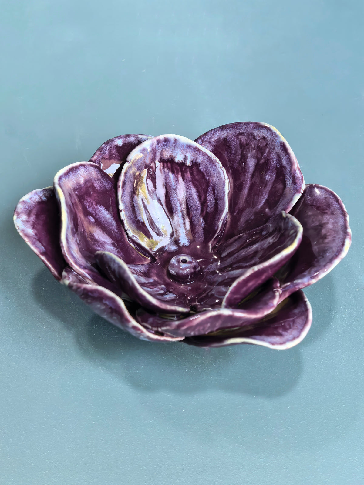 A sculptural ceramic flower bowl in deep purple with glossy highlights and a white glazed edge, set against a solid teal-blue background.