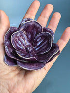 A hand holding a small sculptural flower-shaped dish or bowl in deep purple with glossy white glaze accents, set against a soft blue background.