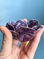A hand holding a small, flower-shaped ceramic bowl with glossy purple petals and white marbled highlights, set against a soft blue background.