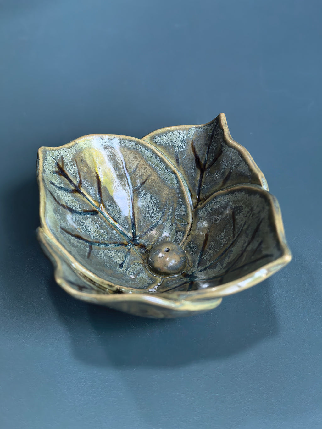 A ceramic leaf-shaped bowl with a glossy, metallic bronze and gold glaze, featuring detailed leaf veins and a small central protruding bead, set against a dark slate-blue background.