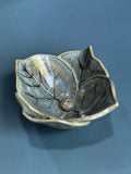 A ceramic leaf-shaped bowl with a glossy, metallic bronze and gold glaze, featuring detailed leaf veins and a small central protruding bead, set against a dark slate-blue background.