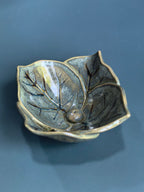 A ceramic leaf-shaped bowl with a glossy, metallic bronze and gold glaze, featuring detailed leaf veins and a small central protruding bead, set against a dark slate-blue background.