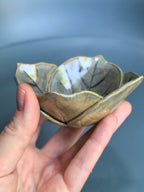A handmade ceramic lotus-shaped bowl with a leaf pattern, held in a person's hand against a soft blue-gray background.