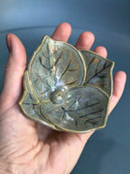 A handmade ceramic leaf-shaped bowl with a glossy olive-green and brown glaze, held in a person's hand against a dark blue-gray background.